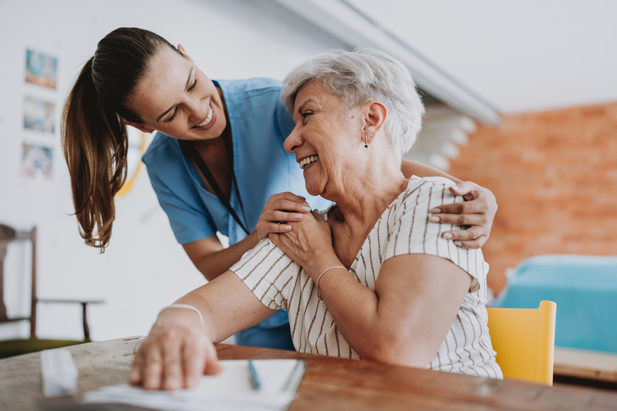 A smiling nurse in blue scrubs gently embraces an elderly woman with gray hair, who is laughing at a table with a notepad in front of her in a memory care community. They appear to be sharing a joyful moment in a bright, cozy room.